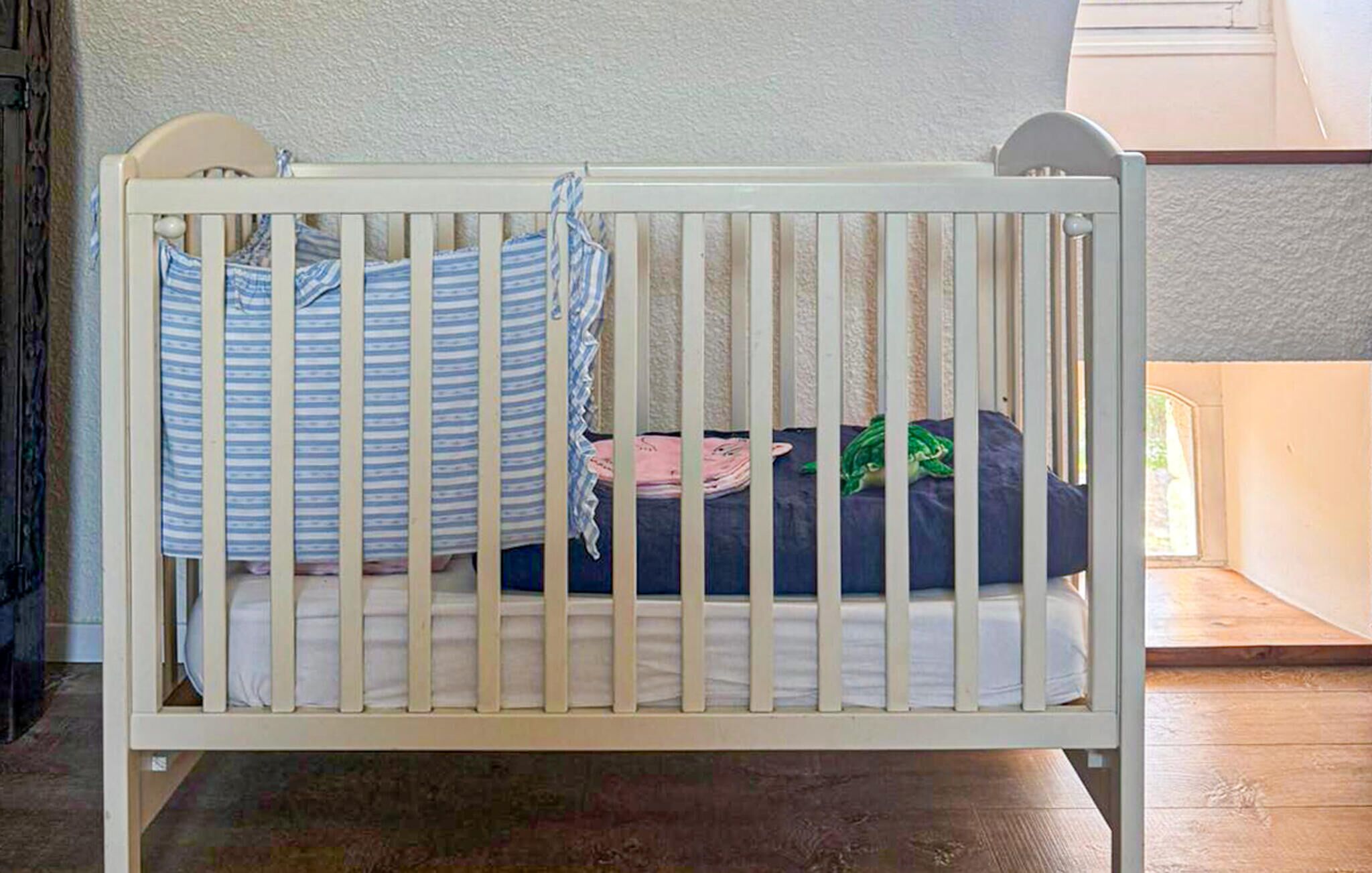 A white crib with a striped pillow and a green blanket, positioned in a room with a wooden floor and a window.