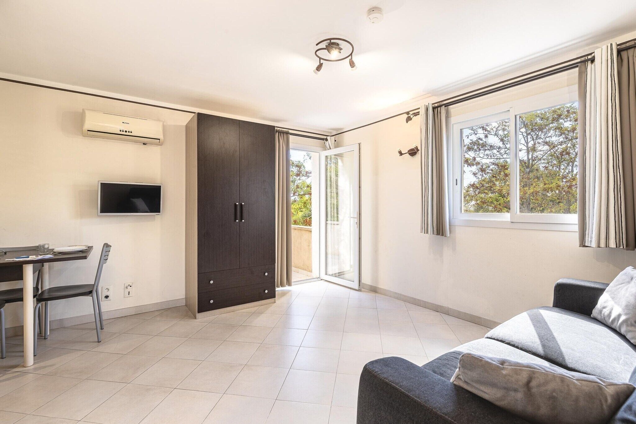 Uma sala de estar moderna com sofá, televisão, área de jantar com mesa e cadeiras, e porta de vidro deslizante que dá para uma área externa.