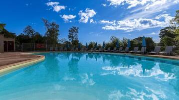 Piscina límpida com espreguiçadeiras e guarda-sóis, cercada por árvores e um céu azul.