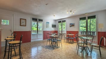 A spacious room with multiple tables and chairs, large windows with a view of greenery, and a television mounted on the wall.