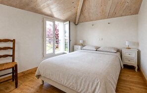 Een slaapkamer met een houten plafond, een groot bed, twee nachtkastjes met lampen, een houten stoel en een raam met uitzicht op bomen.