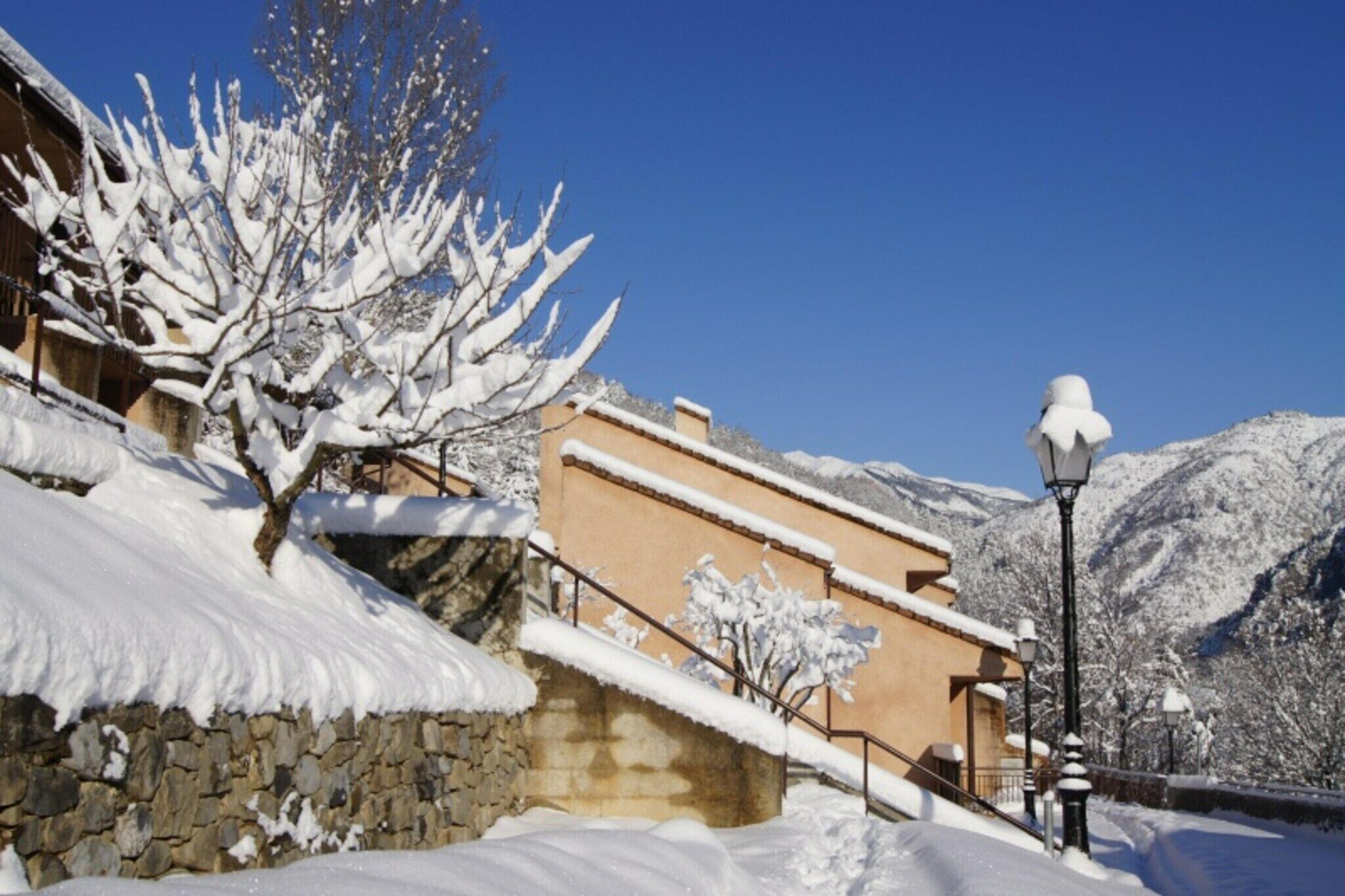 A snow-covered building with a stone wall, a staircase, and a street lamp.
