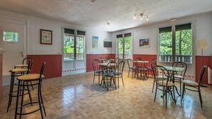 Um quarto amplo com várias mesas e cadeiras, janelas grandes com vista para vegetação, e uma televisão presa na parede.