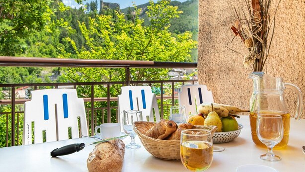 Uma mesa com pão, um jarro de chá e uma cesta de frutas numa varanda com vista para o verde e um prédio.