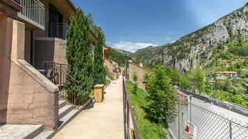 A residential building with a staircase leading to a balcony, surrounded by greenery and mountains in the background.
