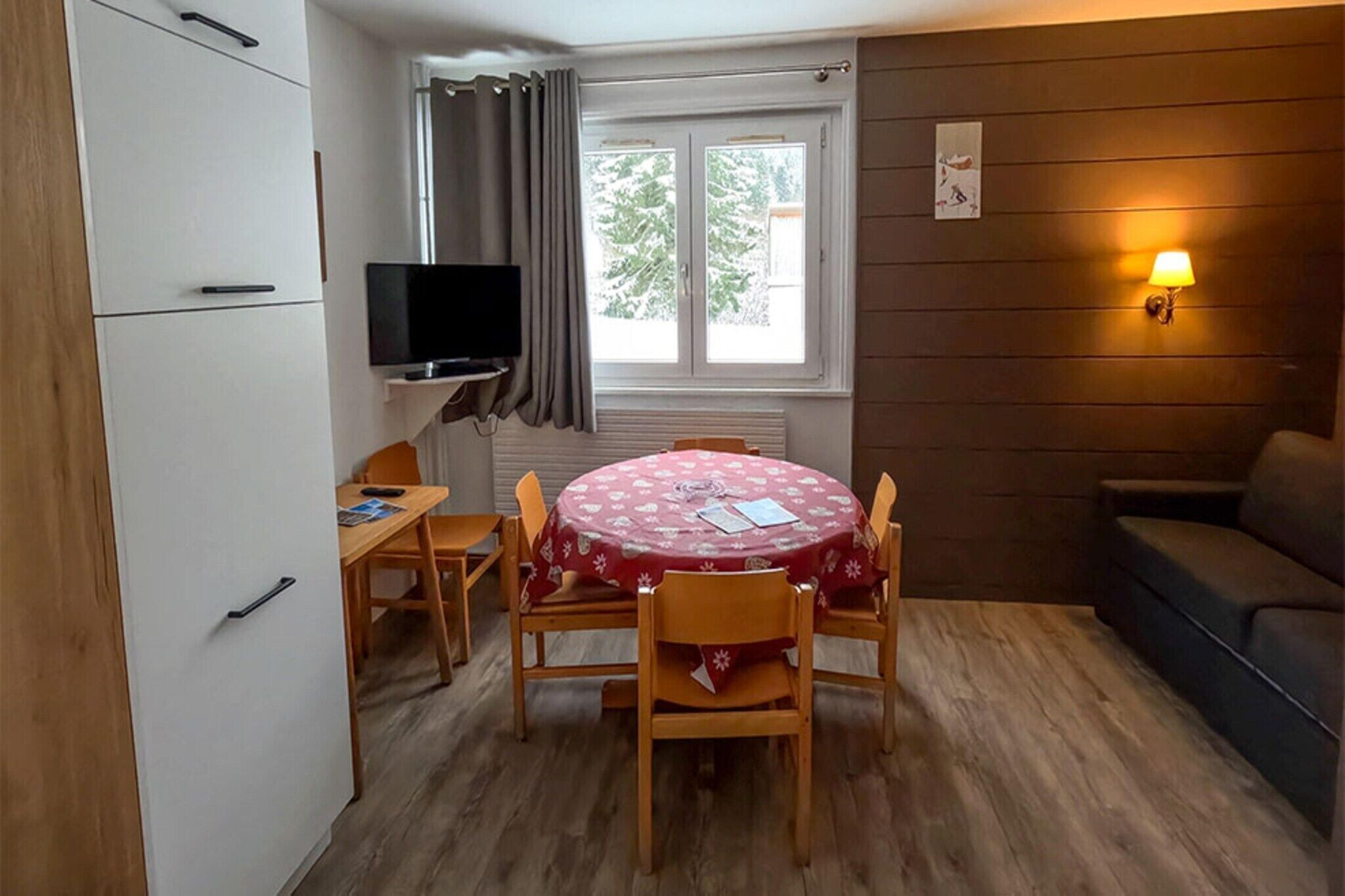 A compact room with a dining table, chairs, a television, a sofa, and a window with curtains.