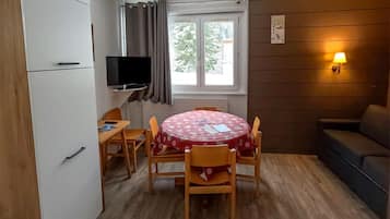 A compact room with a dining table, chairs, a television, a sofa, and a window with curtains.