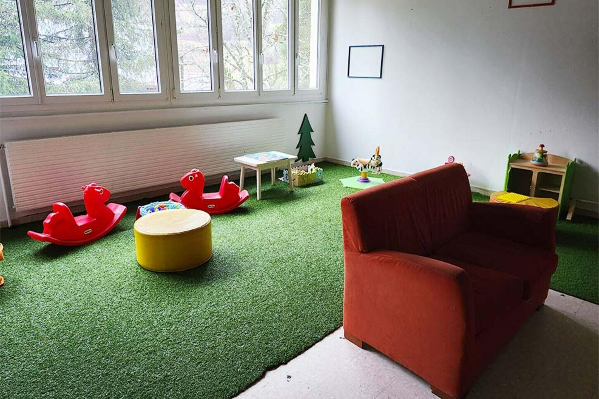 A room with a red sofa, a small table, a green play area, and a window with a view of trees.