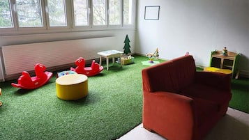 A room with a red sofa, a small table, a green play area, and a window with a view of trees.