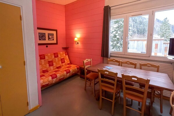 Huoneessa on puinen ruokapöytä, kuusi tuolia, peitolla varustettu sohva, seinään kiinnitetty lamppu ja ikkuna, josta avautuu näkymä puihin.