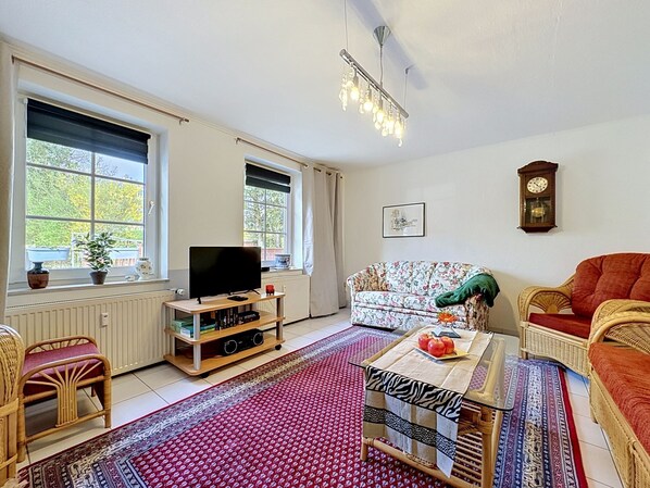Un salon avec une télévision, un canapé, des fauteuils, une table avec des fruits, une horloge et de grandes fenêtres.