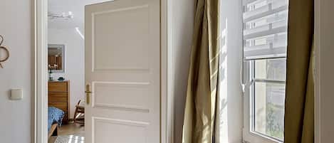 A room with a white door, a radiator, a window with curtains, and a glimpse of a bed with a blue cover.