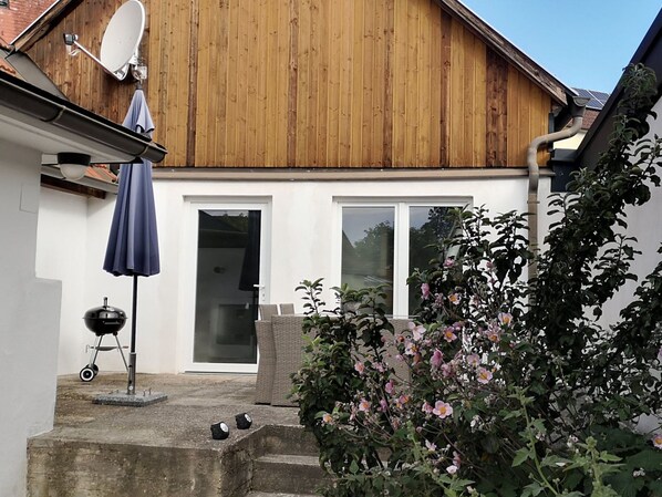 Une maison en bois avec une antenne parabolique, un barbecue et un jardin où poussent des fleurs roses.