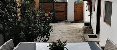Un patio con una mesa y sillas, una planta en maceta, un portón de madera y un edificio blanco con una ventana.