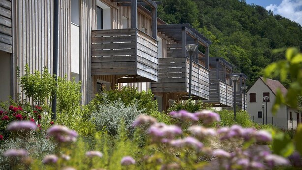 A modern building with a wooden facade, surrounded by lush greenery and flowering plants. - Noir Holiday Apartment for 4 (LES  EYZIES  DE  TAYAC)