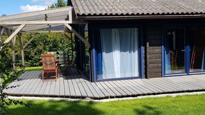 Una terraza de madera con zona de estar cubierta, un edificio con techo de tejas y una puerta de cristal.