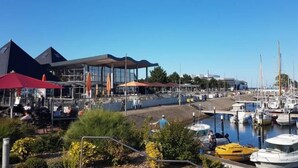 Una marina con barcos, un edificio moderno de fachada de vidrio y zonas de estar al aire libre.
