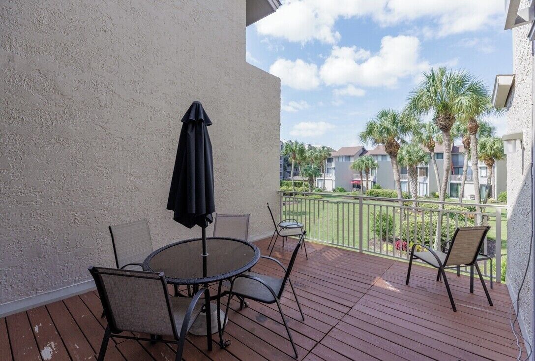 Een balkon met een ronde tafel en stoelen, een zwarte parasol en uitzicht op palmbomen en gebouwen.