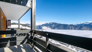 Blick von einem Balkon mit Metallgeländer auf eine Straße mit Fahrzeugen und schneebedeckte Berge unter blauem Himmel.