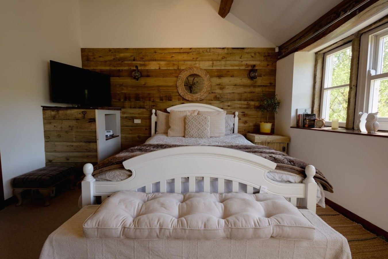 Une chambre avec un mur en bois, un lit avec une tête de lit, un banc, une télévision et une fenêtre avec des rideaux.