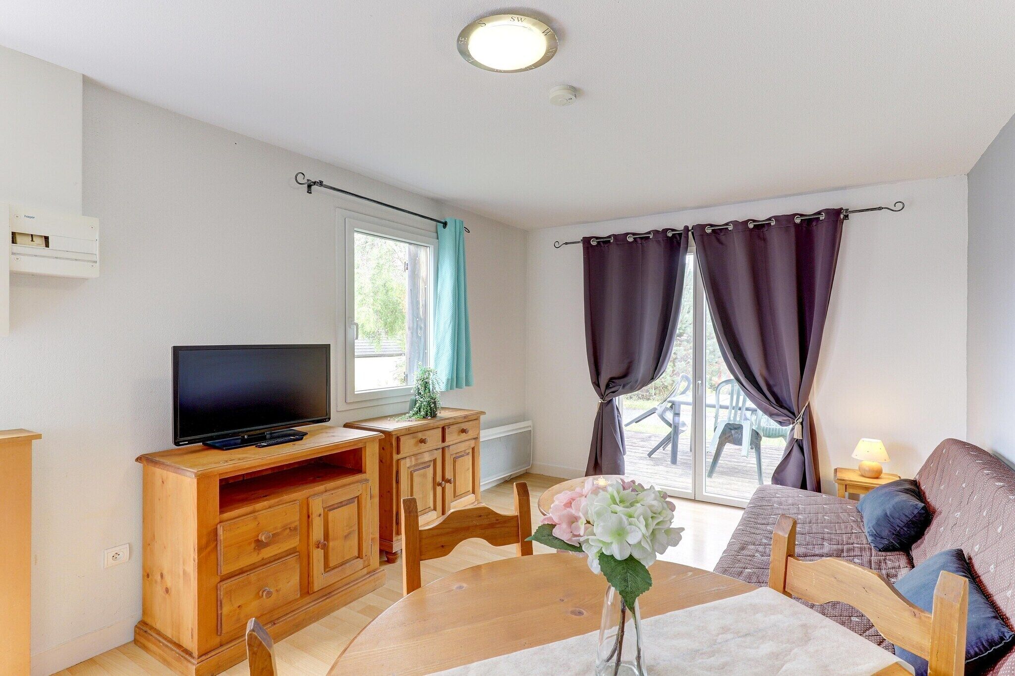 A room with a television on a wooden cabinet, a sofa, a dining table with a vase of flowers, and a window with curtains.