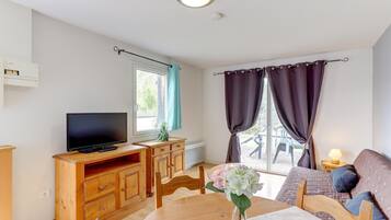 A room with a television on a wooden cabinet, a sofa, a dining table with a vase of flowers, and a window with curtains.