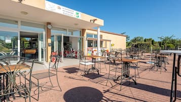A building with a sign reading 'LA GUINGUETTE DES DEMEURES' and a clock. There are outdoor tables and chairs, and a paved area.