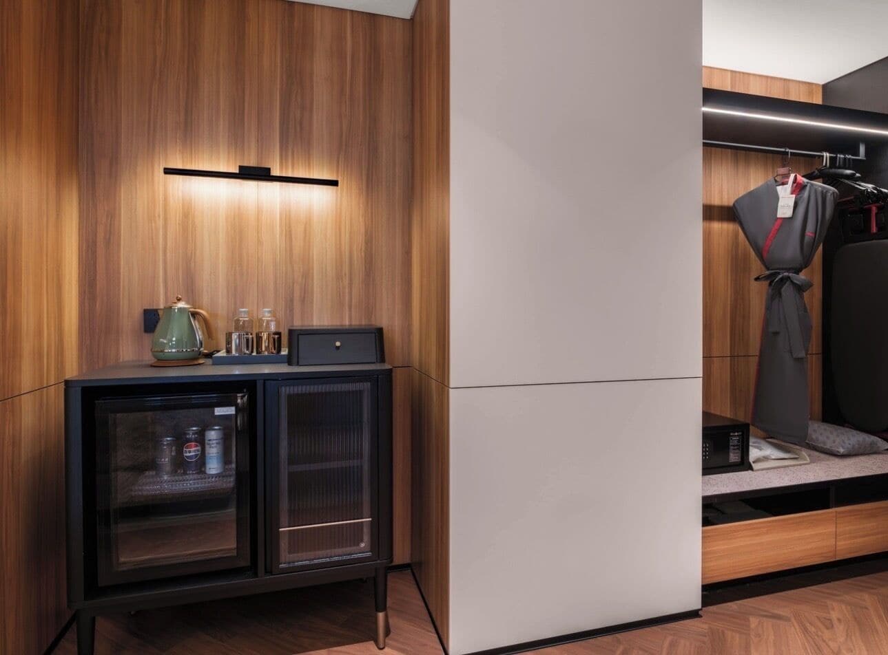 A modern hotel room with a wooden wall, a minibar, a seating area with a sofa and pillows, and a coat rack.
