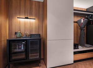 Una habitación de hotel moderna con una pared de madera, un minibar, una zona de estar con sofá y cojines, y un perchero.