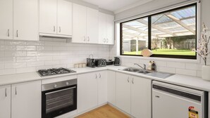 Cozinha moderna com armários brancos, fogão a gás, forno, lava-louças e uma grande janela com vista para um jardim.