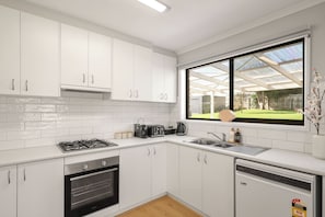 Eine moderne Küche mit weißen Schränken, einem Gasherd, einem Backofen, einem Geschirrspüler und einem großen Fenster mit Gartenblick.