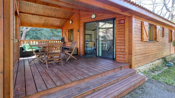 Una cabana de fusta amb un porxo cobert, una zona de dinar amb cadires i una entrada amb porta corredissa de vidre.
