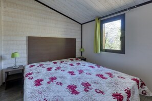 A bedroom with a wooden headboard, a bed with a floral bedspread, a nightstand with a lamp, and a window with a curtain.