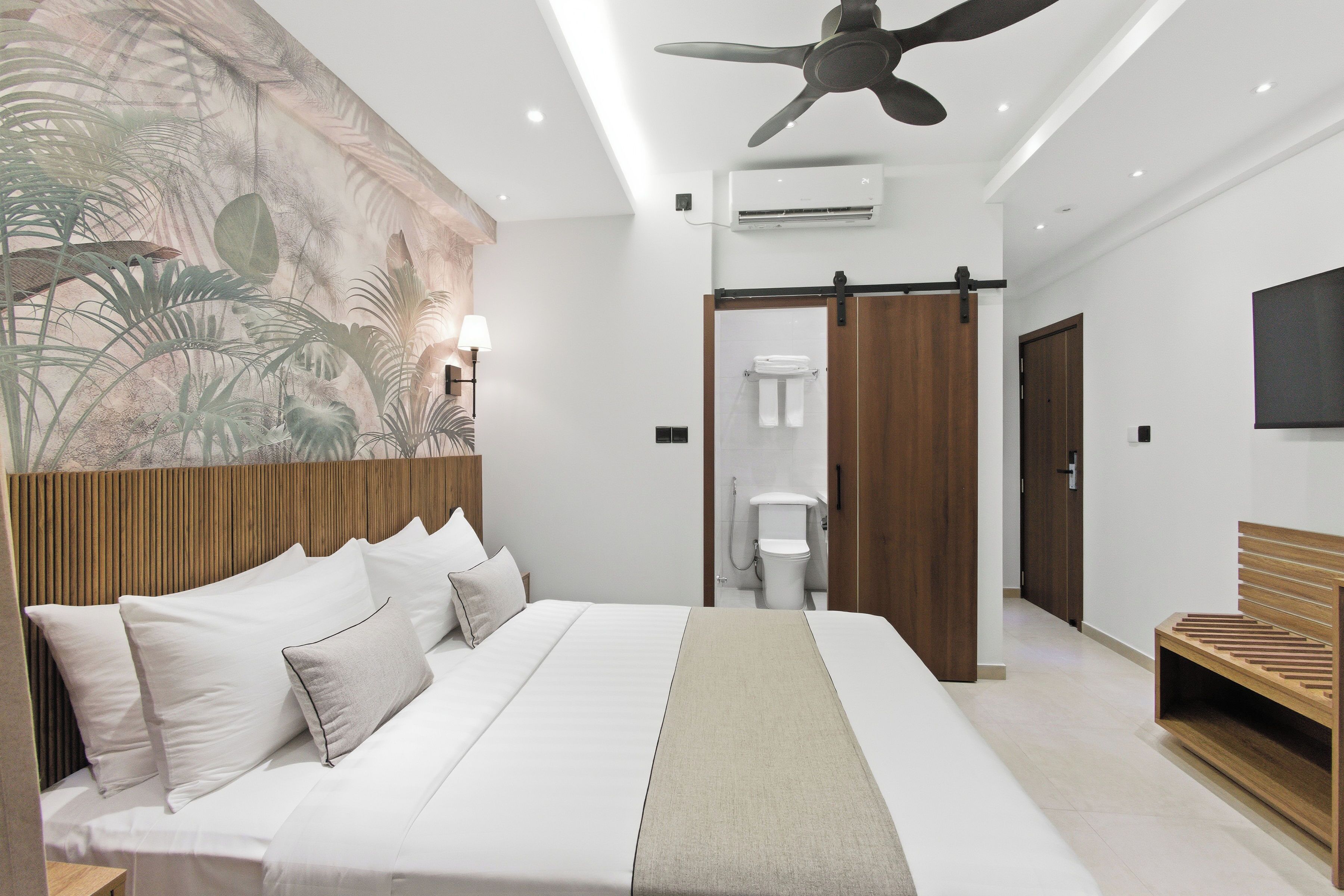 Camera d'albergo moderna con un letto grande, una ventola a soffitto, un condizionatore d'aria, una testiera in legno, una TV fissata al muro e un bagno con il water e il lavandino.