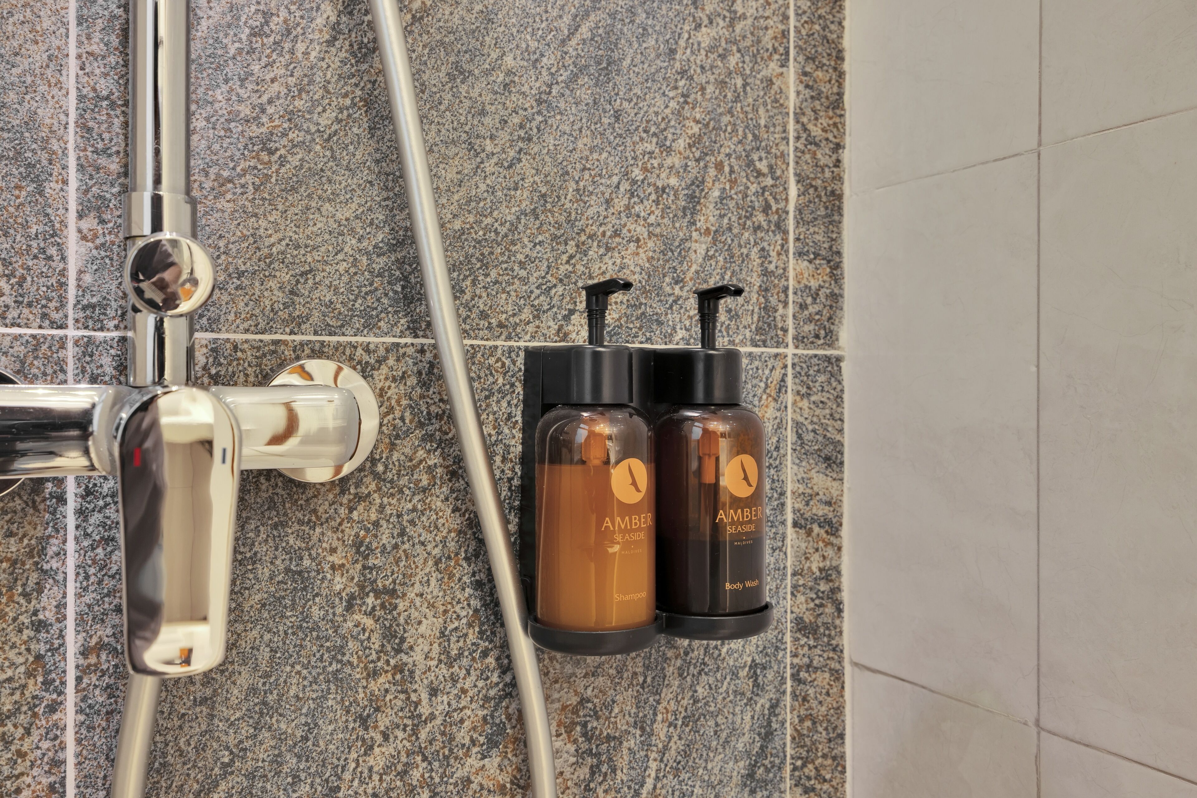 Un área de ducha con grifo cromado, dos botellas color ámbar con la etiqueta 'AMBER SEASIDE' para champú y gel de ducha, y un cabezal de ducha.