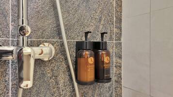 Un área de ducha con grifo cromado, dos botellas color ámbar con la etiqueta 'AMBER SEASIDE' para champú y gel de ducha, y un cabezal de ducha.