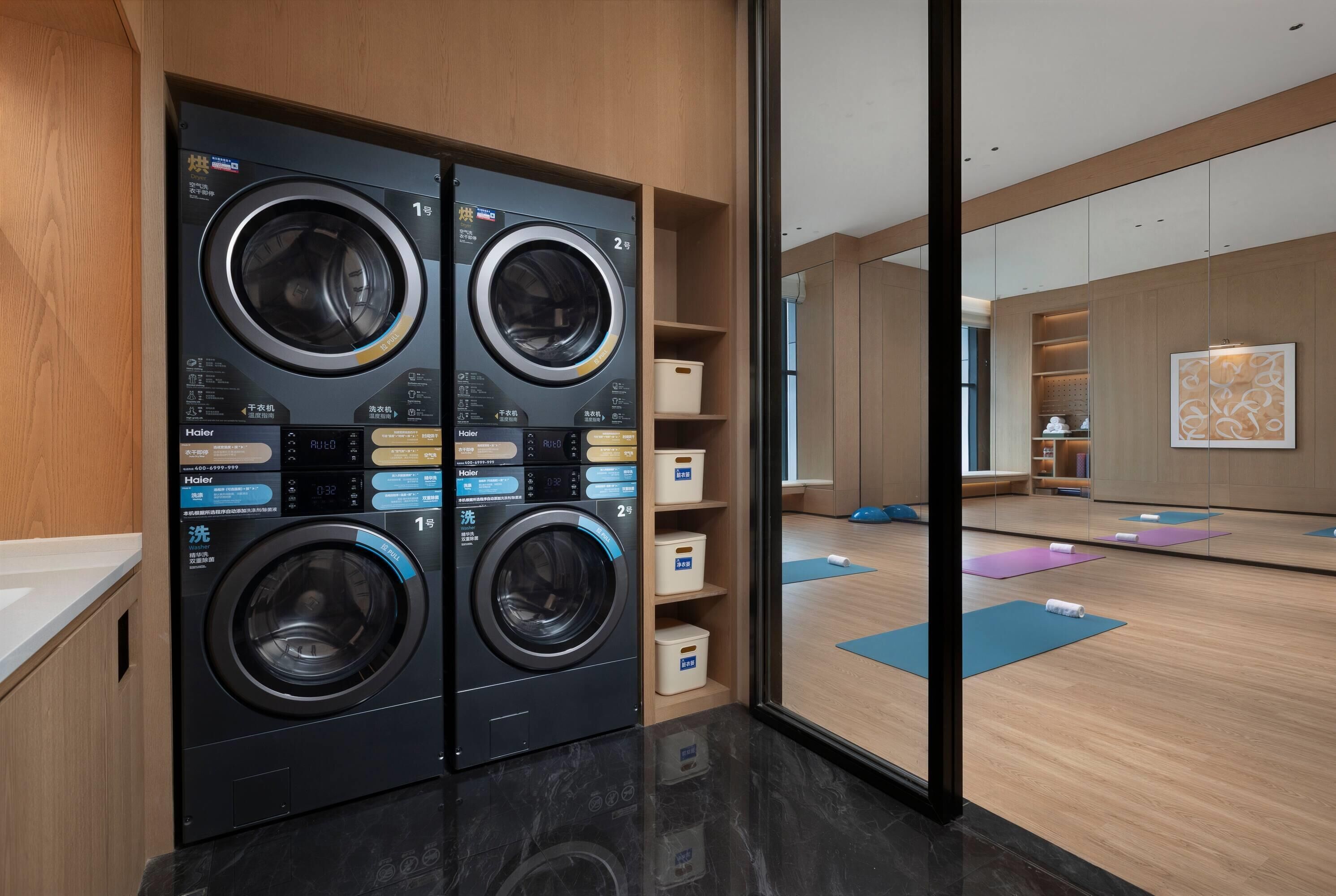Un moderno lavadero con cuatro lavadoras Haier, un mueble de madera y una zona para hacer yoga visible a través de un panel de vidrio.