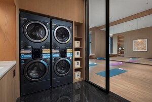 Un moderno lavadero con cuatro lavadoras Haier, un mueble de madera y una zona para hacer yoga visible a través de un panel de vidrio.