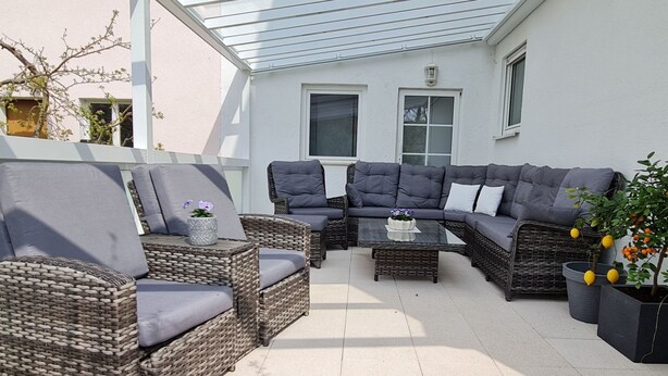 Ein überdachter Patio-Bereich mit Korbmöbeln, einem Glastisch und Blumentöpfen.