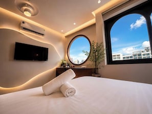 Une chambre d’hôtel moderne avec un grand lit, un téléviseur fixé au mur, un miroir rond et une plante en pot.