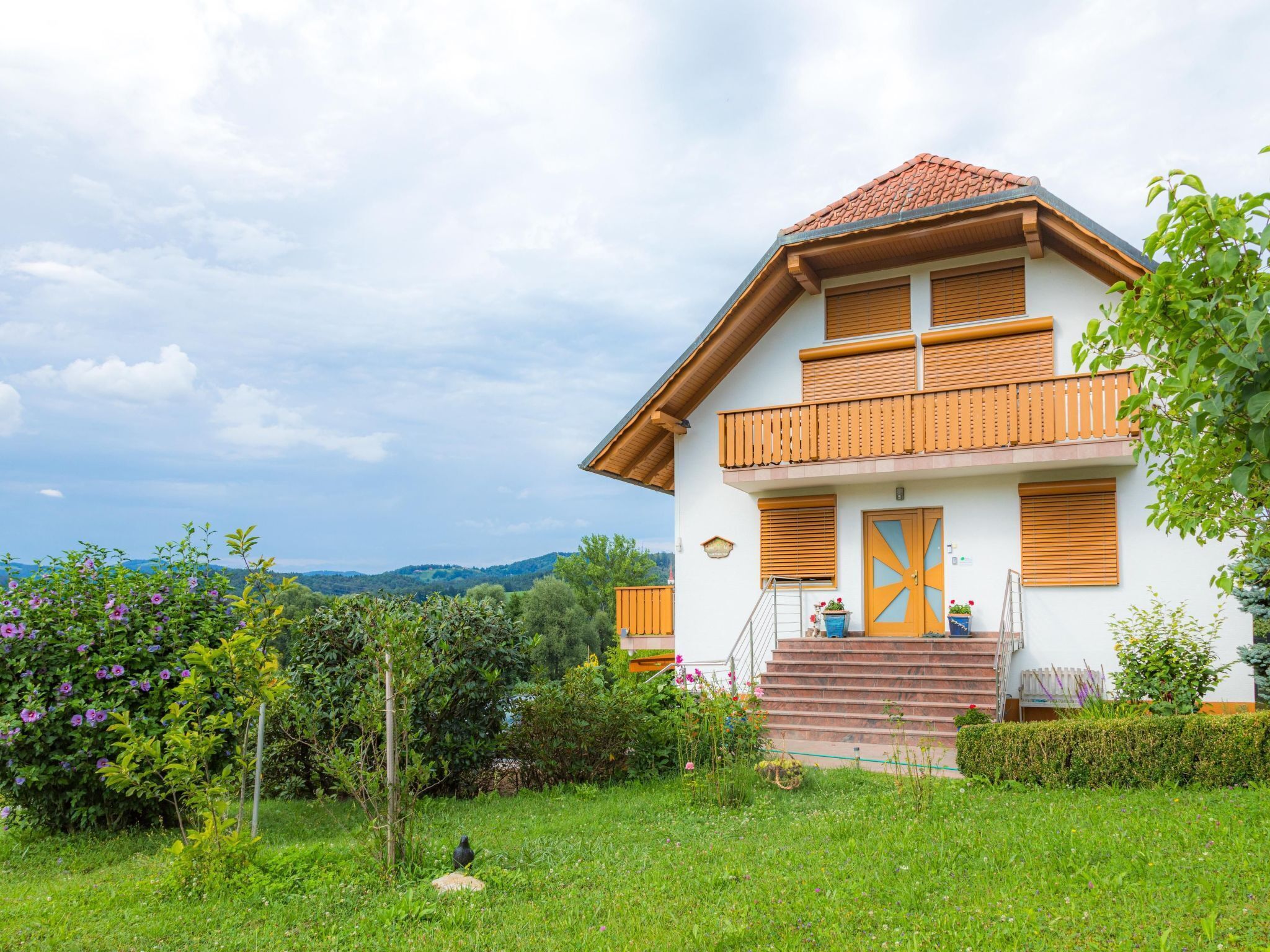 A two-story house with a balcony, a front door with a cross design, and a garden with flowers and greenery.