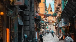 Una calle estrecha bordeada de edificios, algunos con letreros y toldos, que conduce a un edificio lejano con techo puntiagudo.