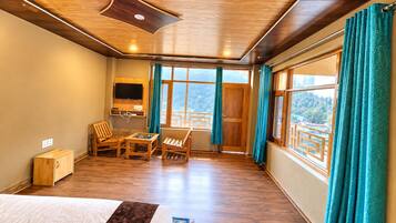 A room with a bed, a TV, a table with chairs, and large windows with curtains.