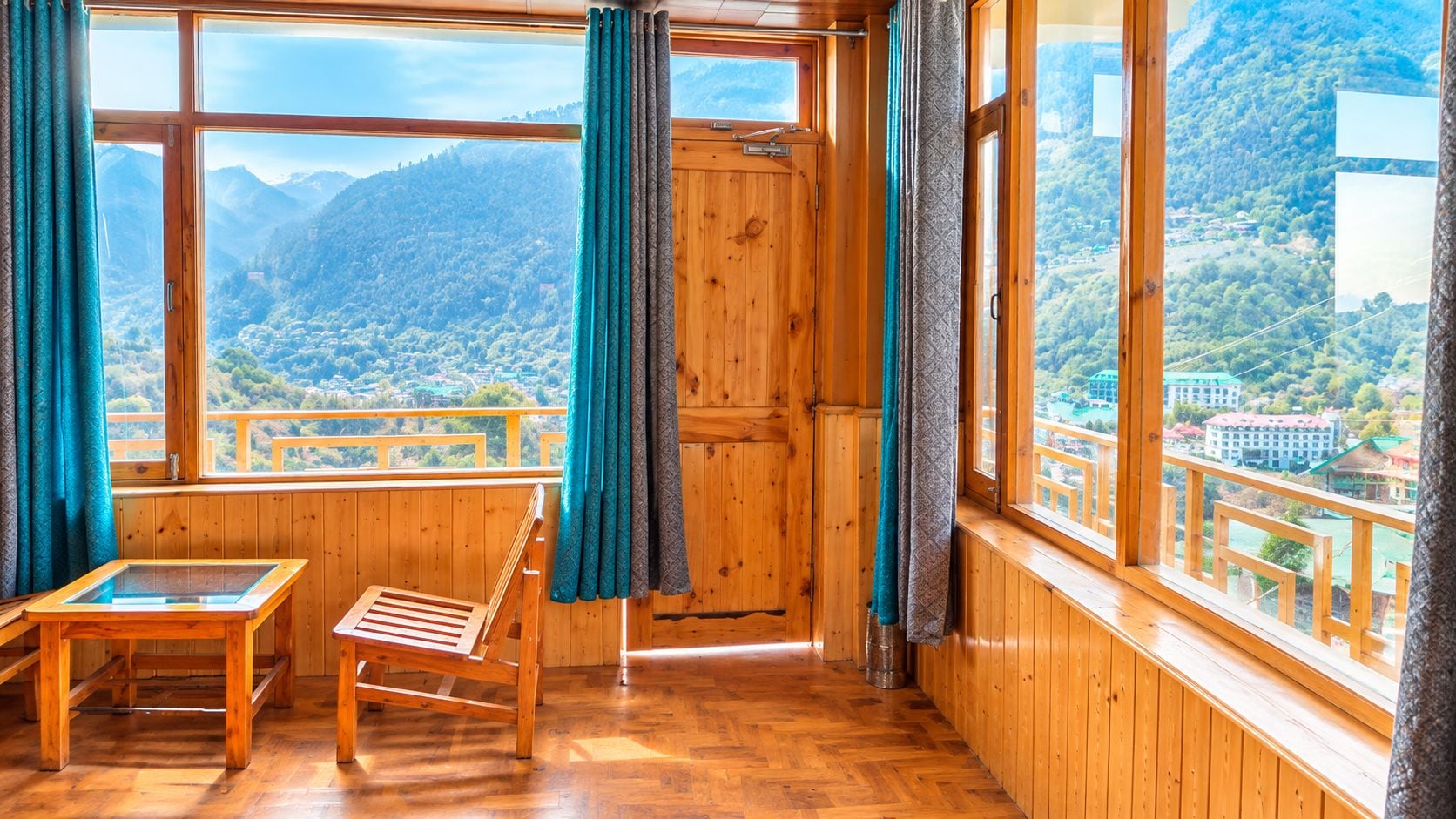 A wooden room with a wooden table and chair, large windows with curtains, and a view of a mountainous landscape.