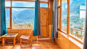 Ein Zimmer mit Holzboden, einem Holztisch und Stuhl, großen Fenstern mit Vorhängen und Blick auf eine gebirgige Landschaft.