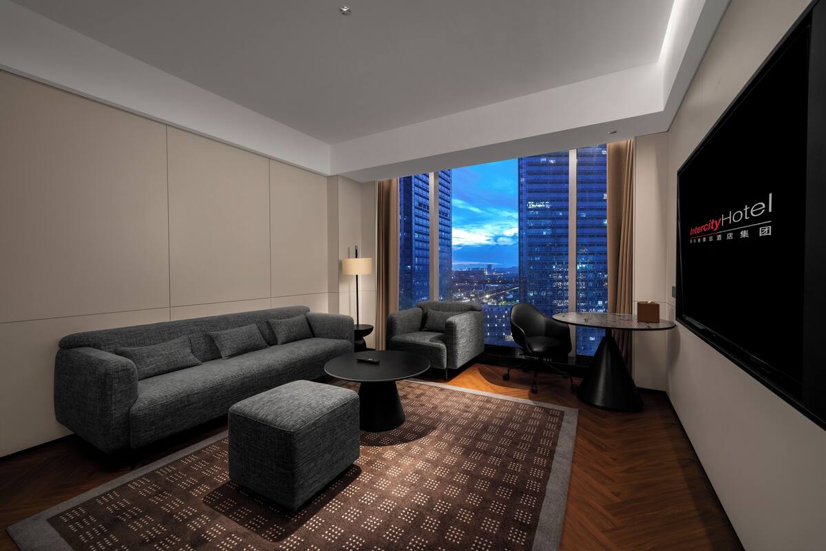 A modern hotel room with a large window, a sofa, a small table, and a TV screen.