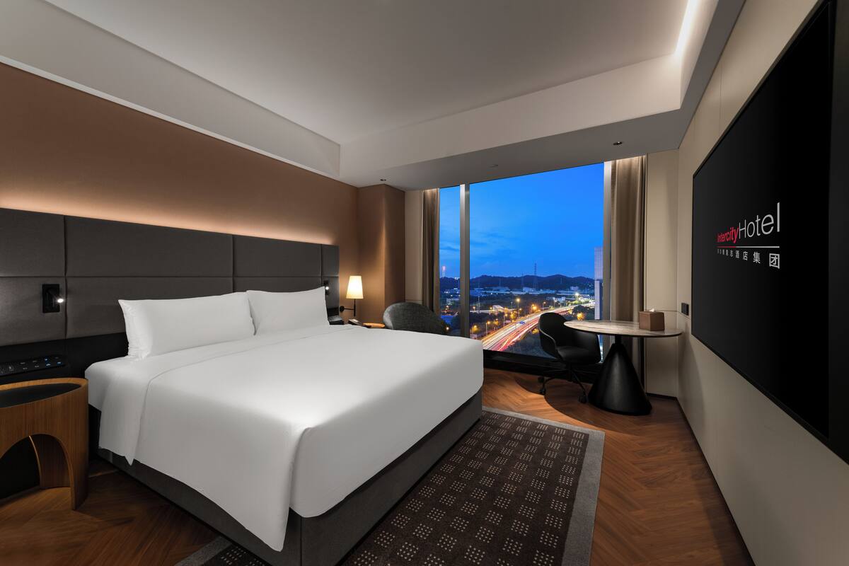 A modern hotel room with a large bed, a desk, and a view of the cityscape.