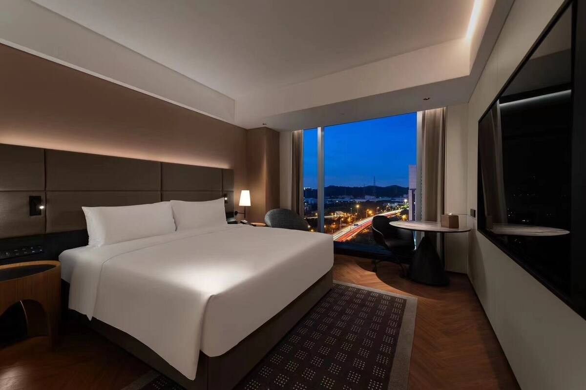 A modern hotel room with a large bed, a desk, a chair, a TV, and a view of a cityscape at dusk.