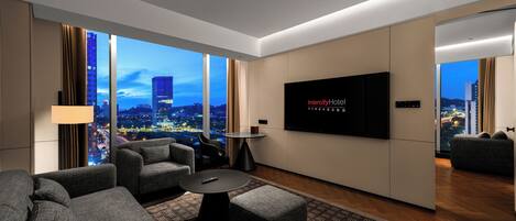 Habitación de hotel moderna con un televisor grande, asientos cómodos, una mesita y vistas a la ciudad.