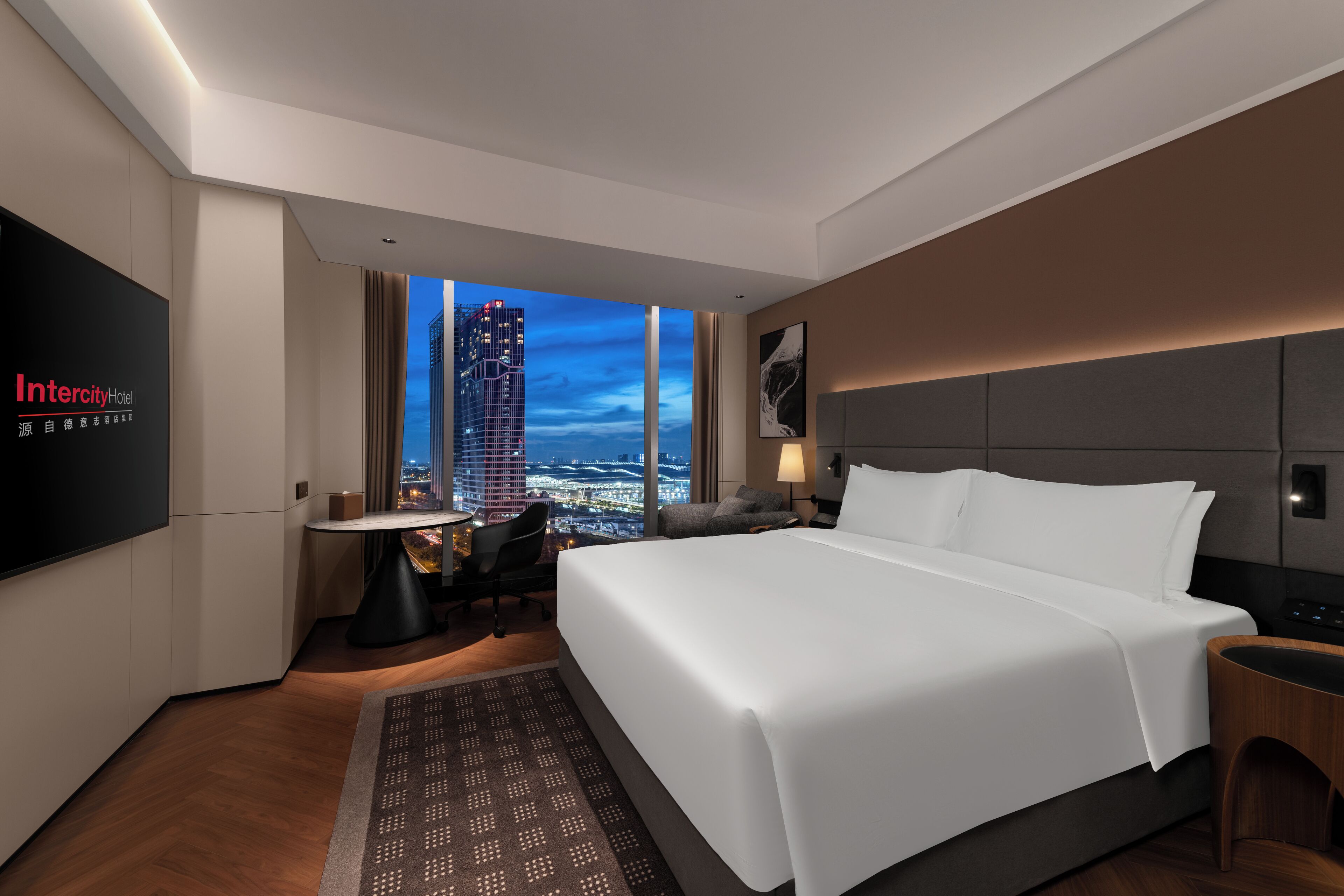 Et moderne hotelværelse med en stor seng, et skrivebord, en stol og et fjernsyn, der viser hotellets navn.